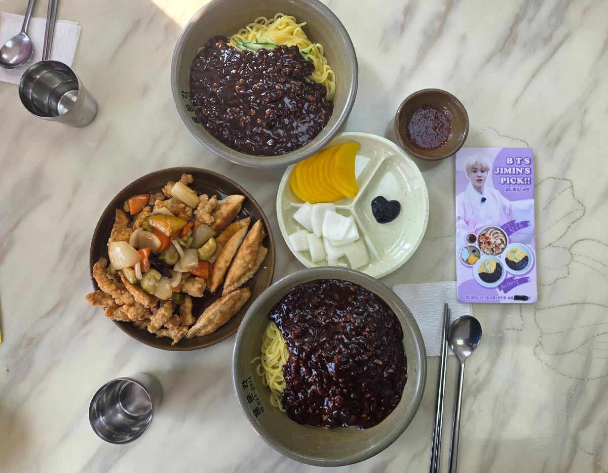 two bowls of jajangmyeon with black bean sauce, fried sweet-and-sour chicken, pickled radish and onions, and BTS-themed pamphlet on marble table