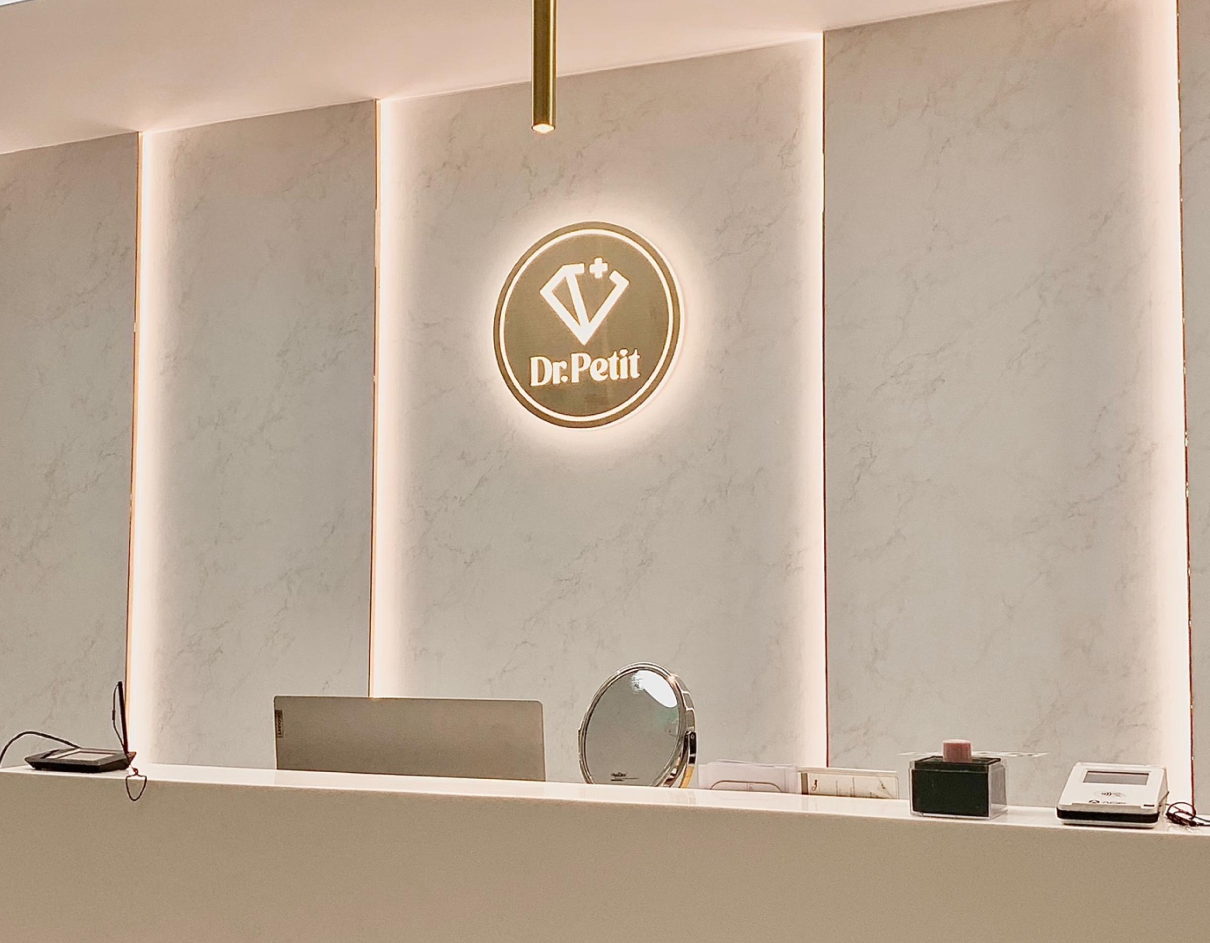 Minimalist white clinic reception desk with illuminated Dr.Petit logo, pendant lights, laptop, and receptionist peeking behind counter