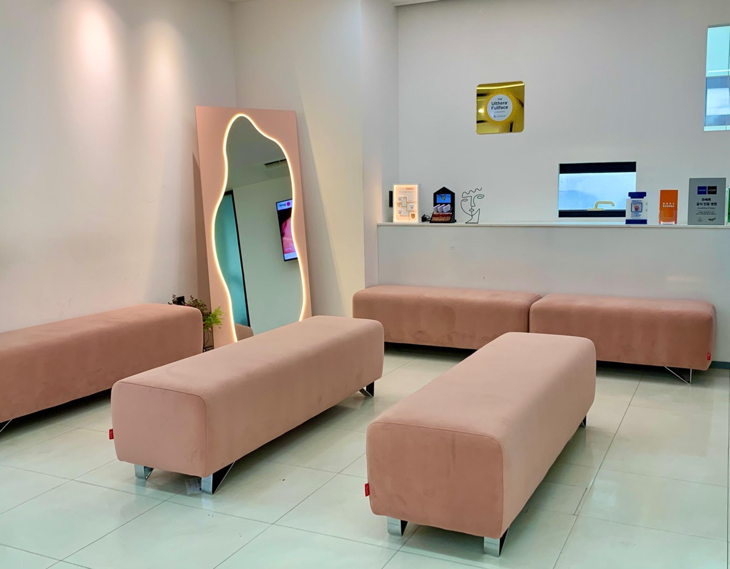 Bright clinic waiting room with pink upholstered benches, illuminated irregular mirror, small shelf with decor and windows