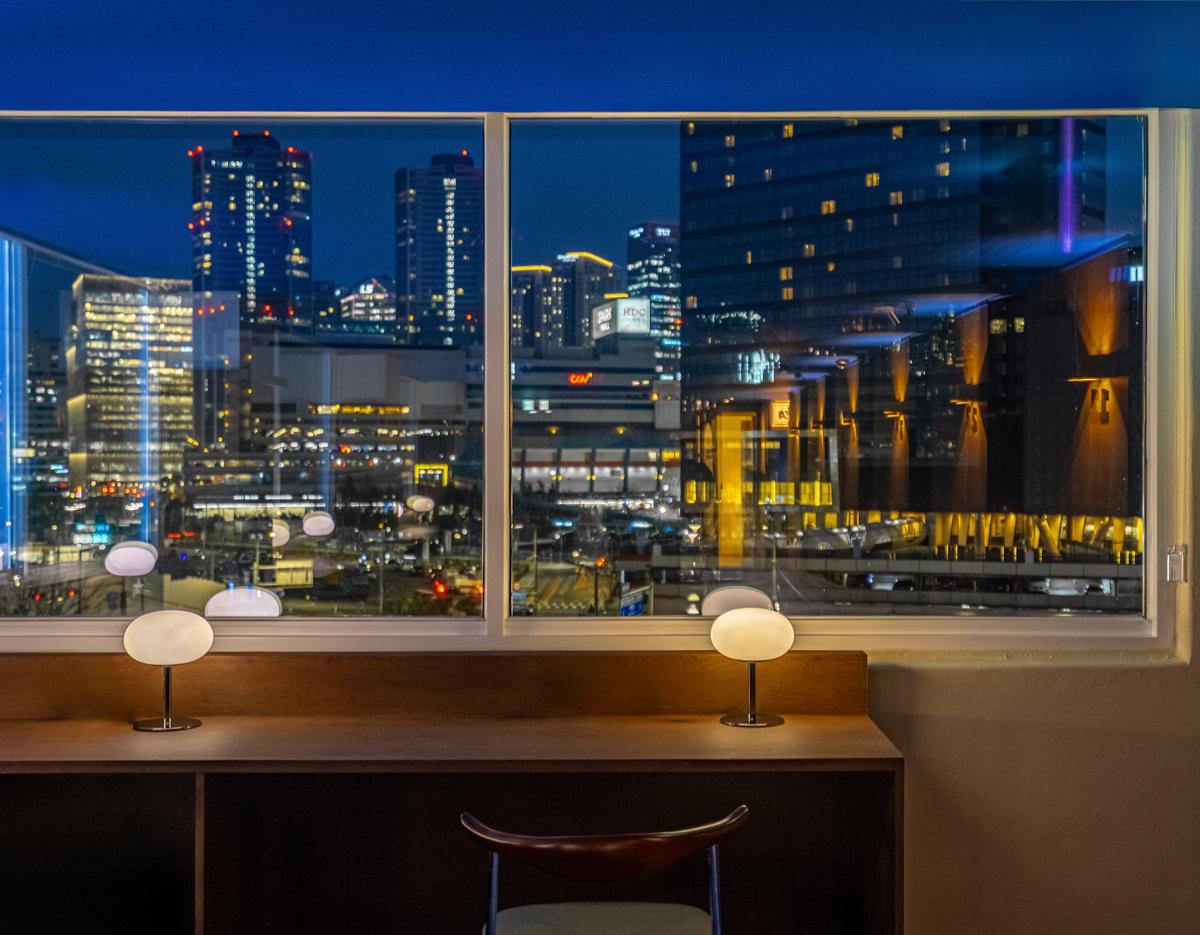 nighttime city skyline seen through lounge window above wooden desk with two modern lamps and a single chair