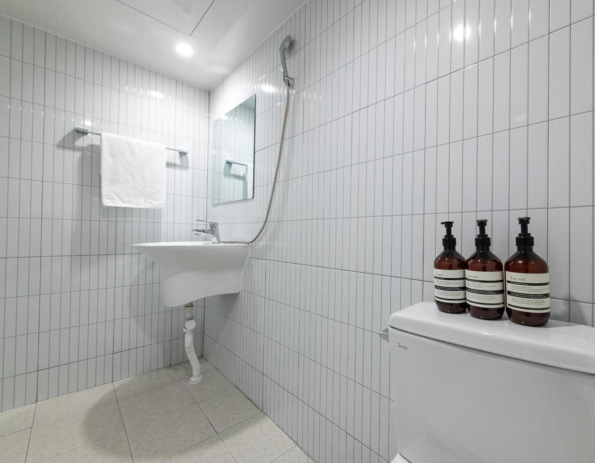 Minimal white tiled guesthouse bathroom with wall-mounted sink, mirror, showerhead, towel and three soap dispensers on toilet