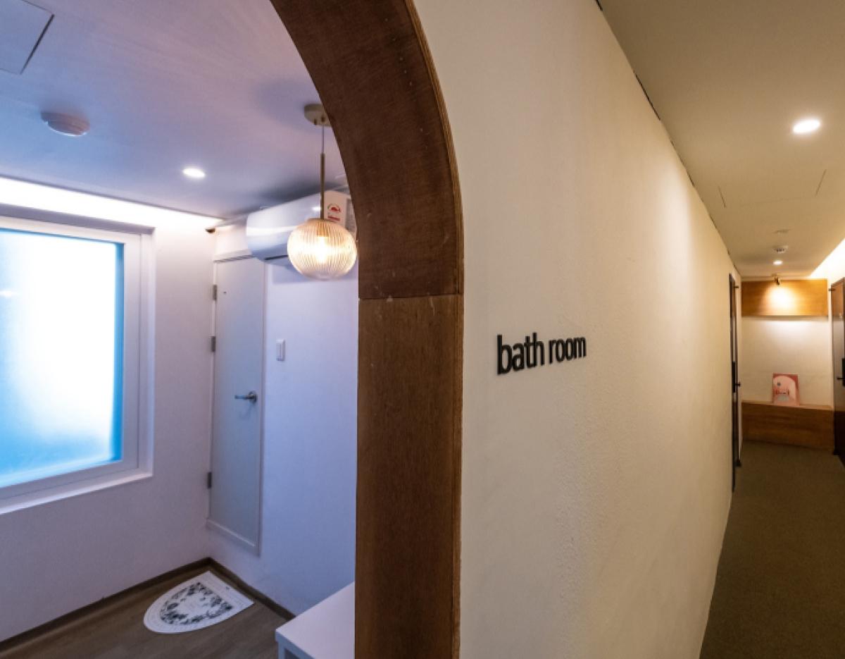 narrow guesthouse corridor with arched wooden doorway and "bath room" sign leading to a softly lit bathroom area