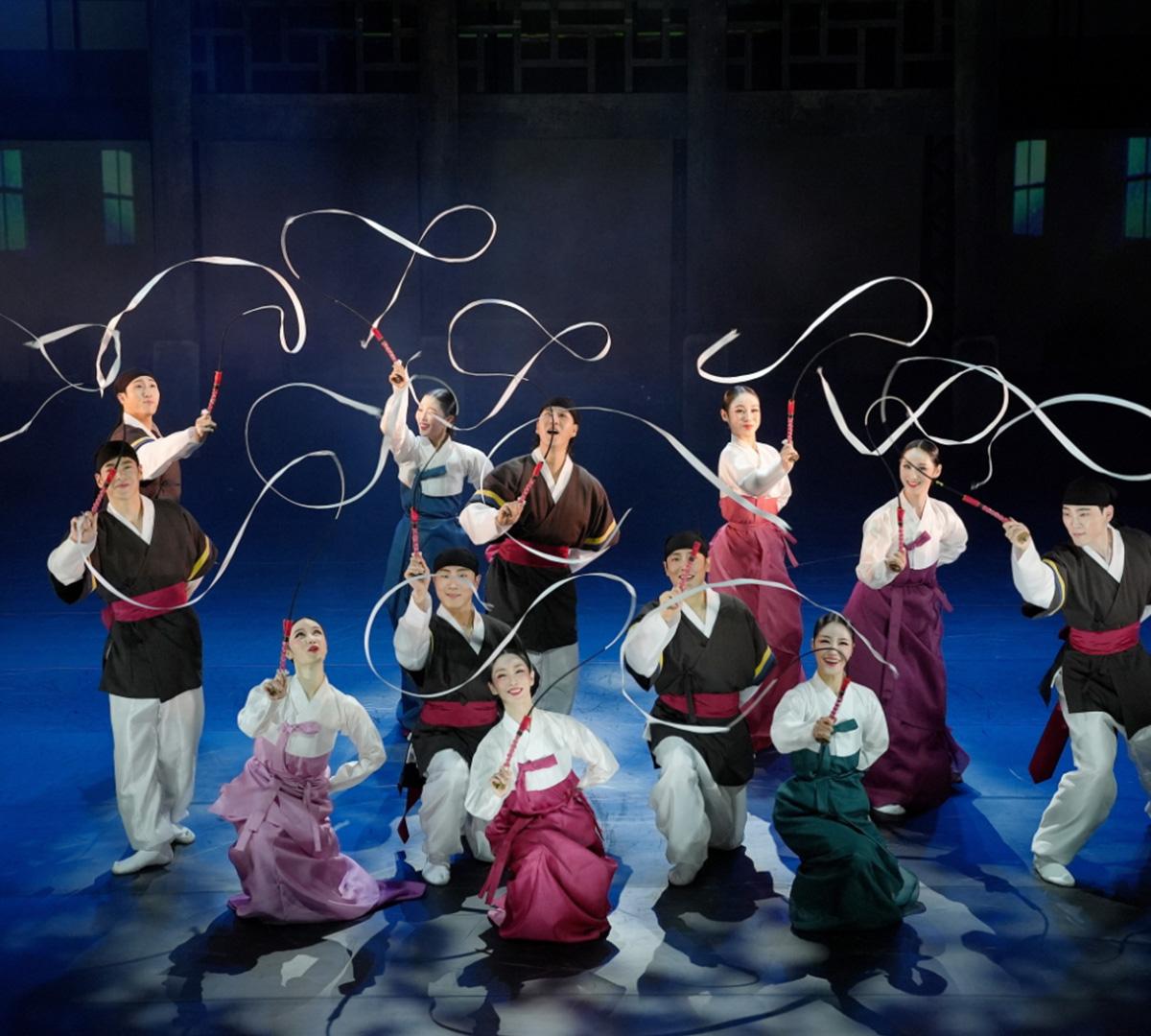 Korean traditional performers in hanbok kneeling and standing on stage, waving long white ribbon streamers from sticks in choreographed formation