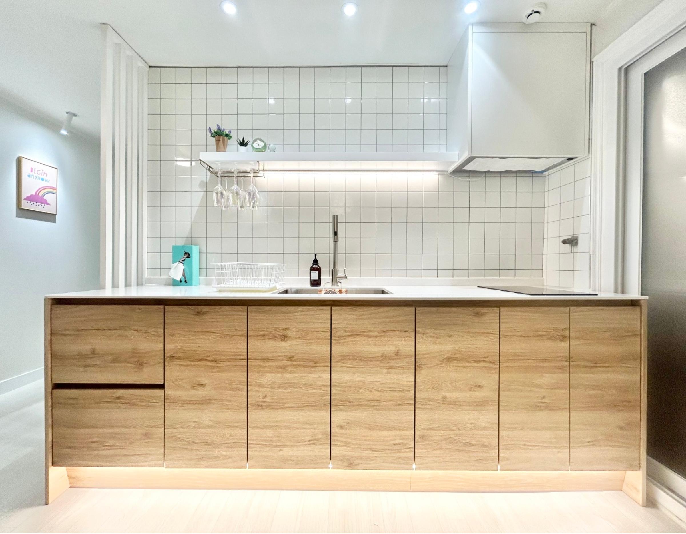 Minimalist kitchen island with wood-grain cabinets, white countertop, sink, tiled backsplash and hanging wine glasses.