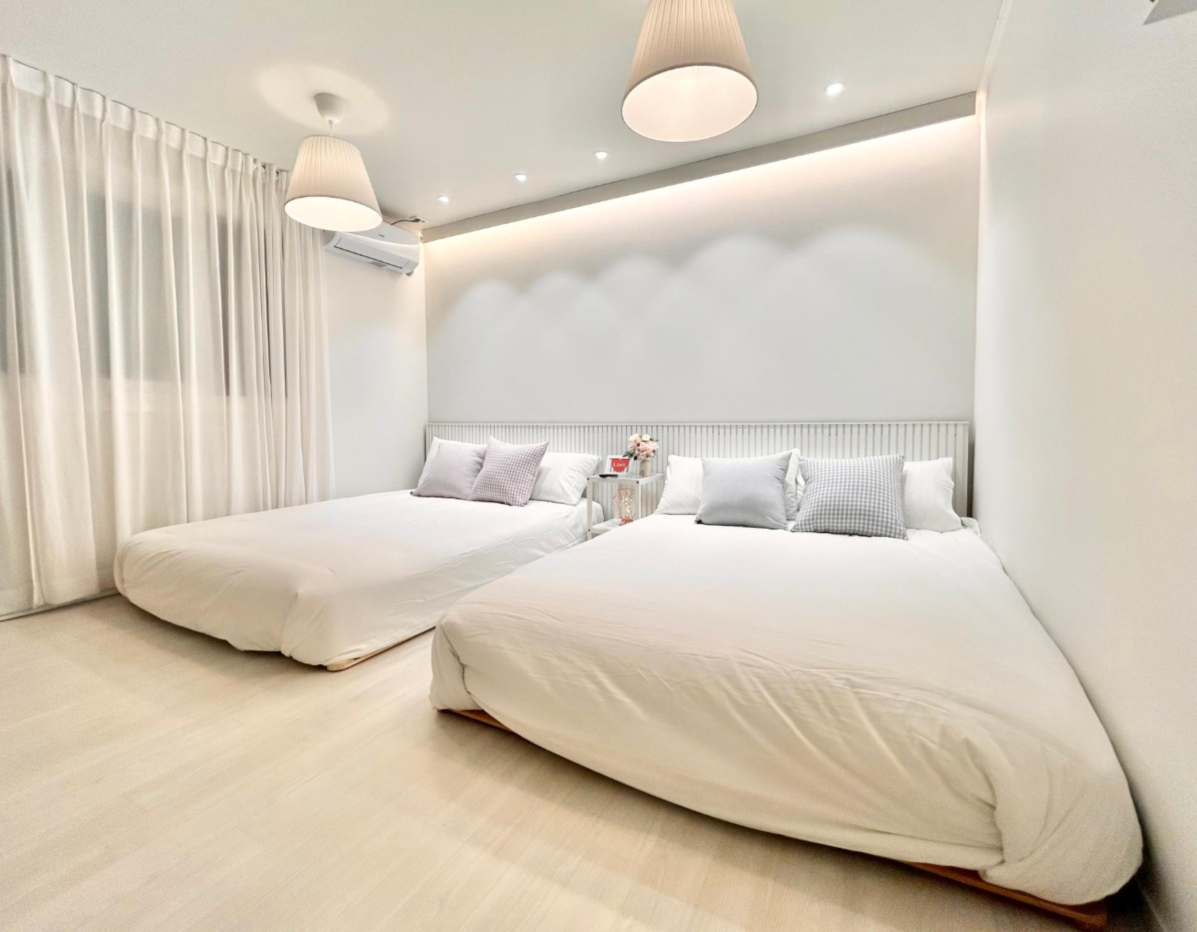 Bright minimalist bedroom with two low queen beds, white bedding, soft pillows, pendant lights and sheer curtains.