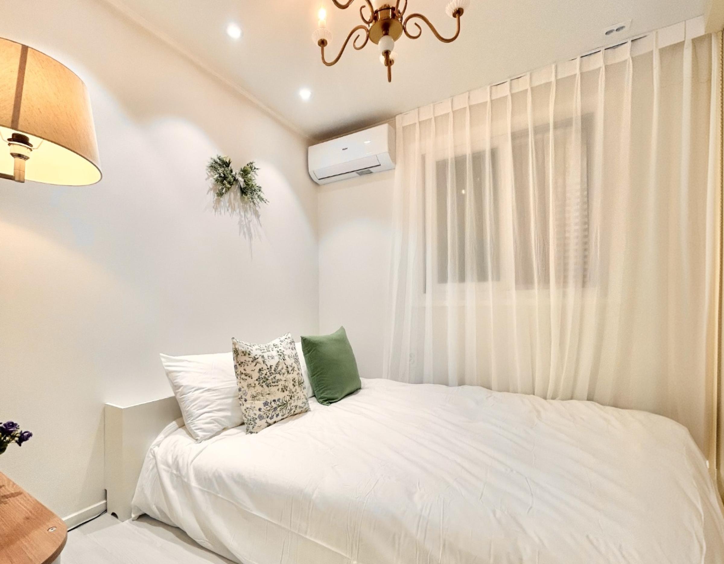 Cozy bedroom with queen bed, white linens, floral and green pillows, sheer curtain, wall-mounted air conditioner and lamp