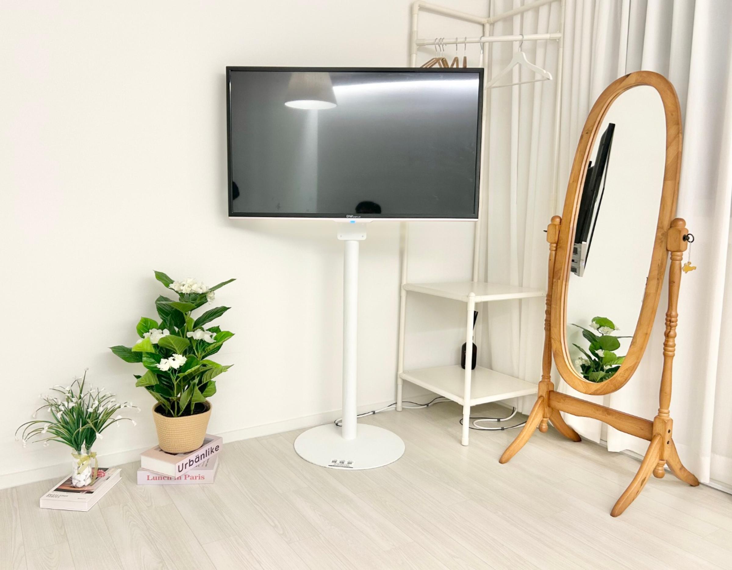 minimal white bedroom corner with freestanding TV on a pedestal, wooden full-length mirror, potted plants and small shelving unit