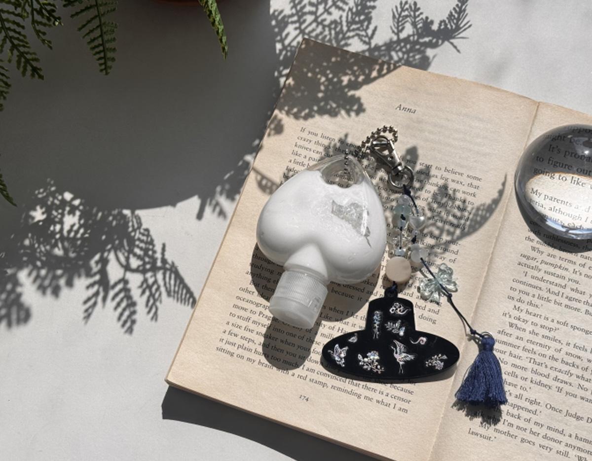 heart-shaped white hand sanitizer bottle and embroidered navy tassel keyring resting on open book pages with fern shadows