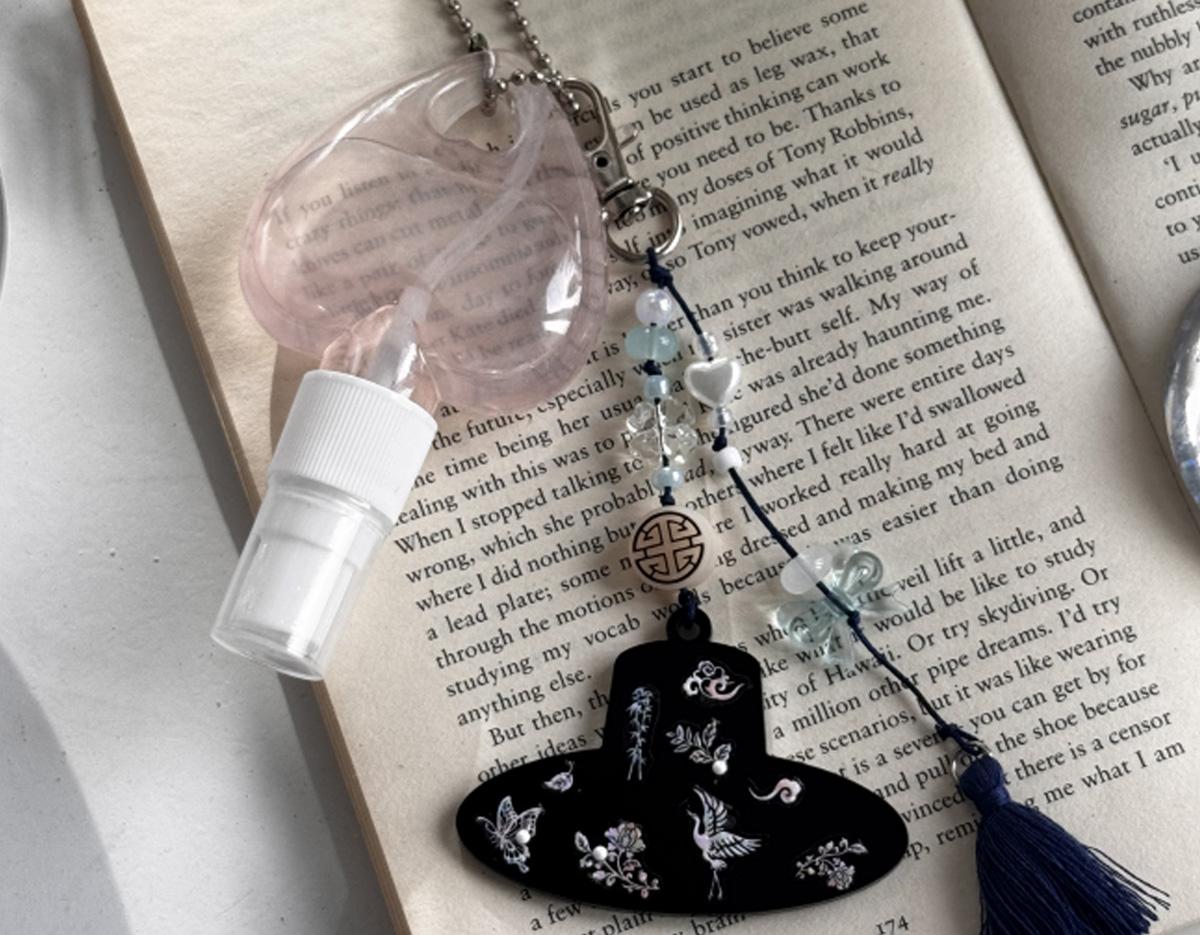 transparent pink heart spray bottle and decorative Korean-style keyring with black hat charm, beads, and blue tassel on open book