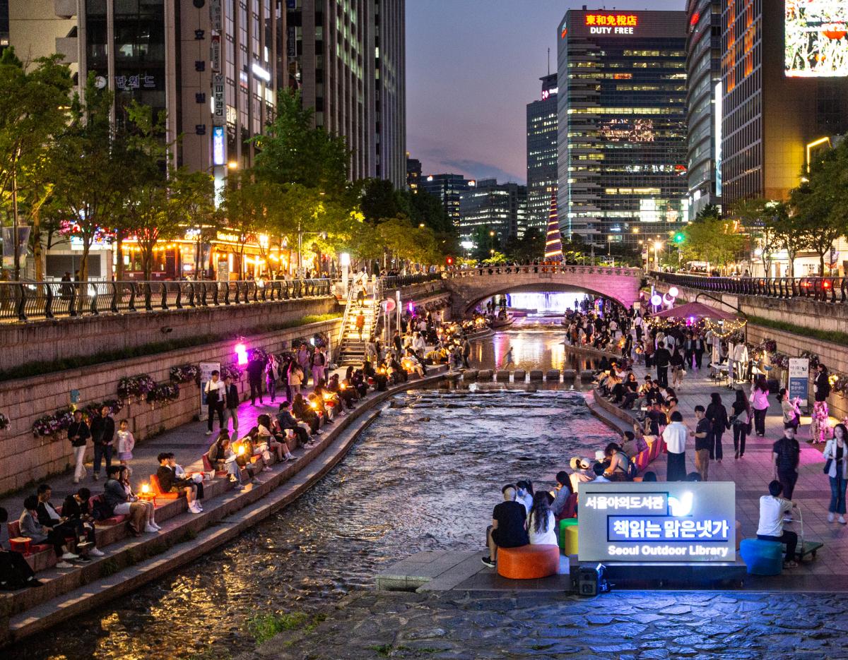Cheonggyecheon stream at night with crowds seated along stepped banks, lit signage reading "Seoul Outdoor Library" and city buildings