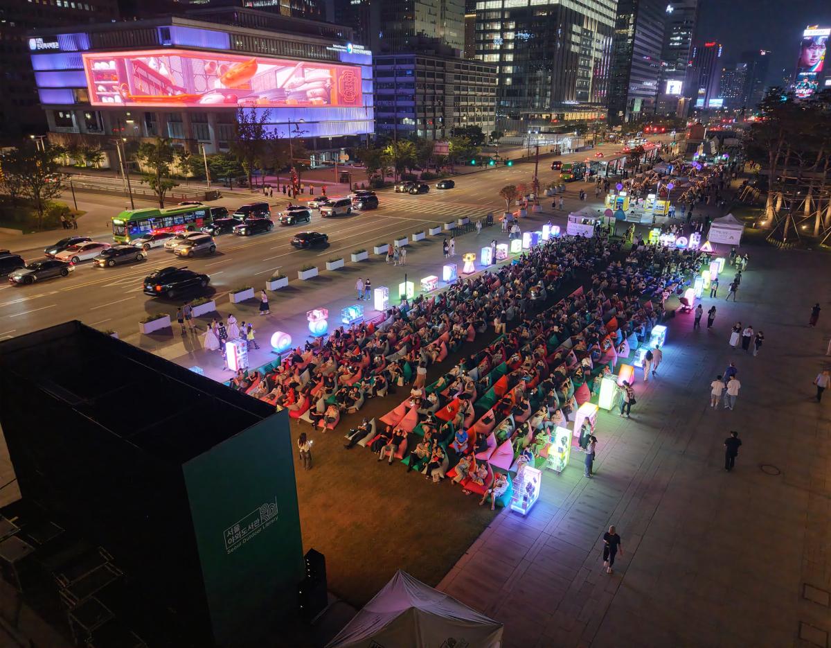 Seoul Plaza at night with rows of people on colorful beanbags and glowing book displays beside a busy city street
