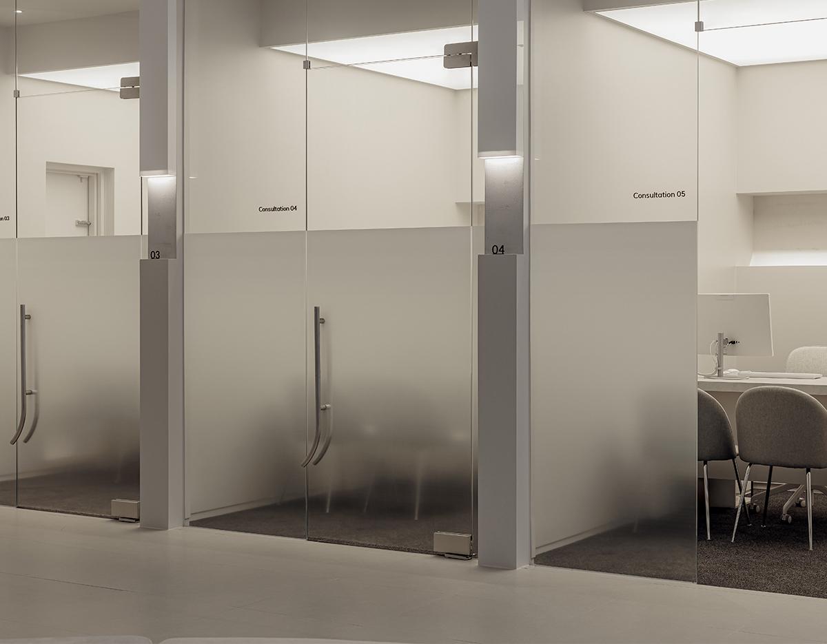 Minimalist clinic hallway with frosted glass consultation rooms numbered 03–05 and chairs visible through glass panels