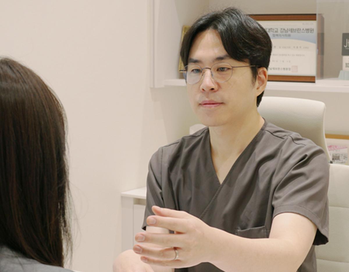 doctor in gray scrubs gesturing with hands while consulting a seated patient in clinic room