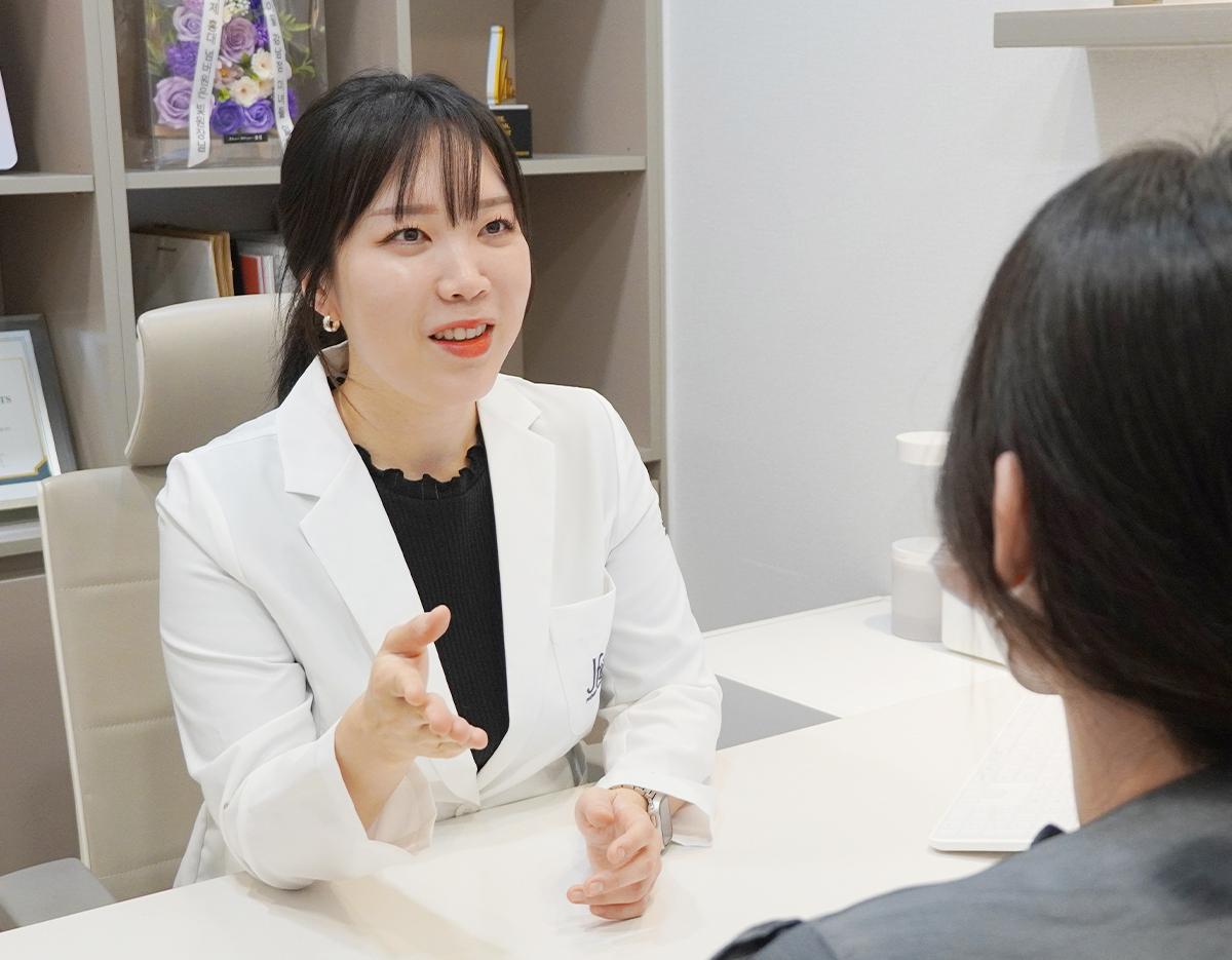Doctor in a white coat gesturing with open hand during a consultation across a desk with a patient visible from behind