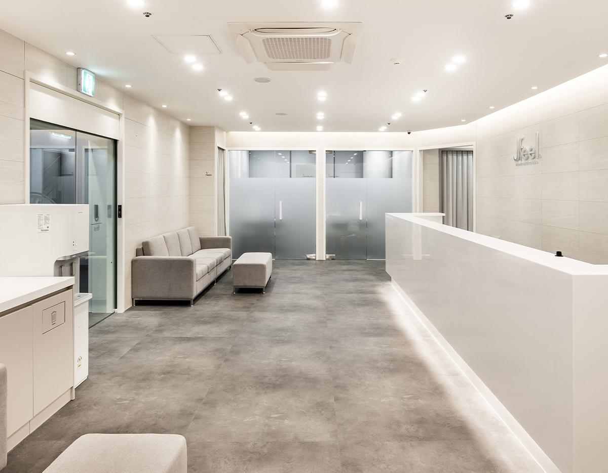 Minimalist bright clinic reception with white desk, gray sofa and frosted glass doors at 제이필의원 홍대점.