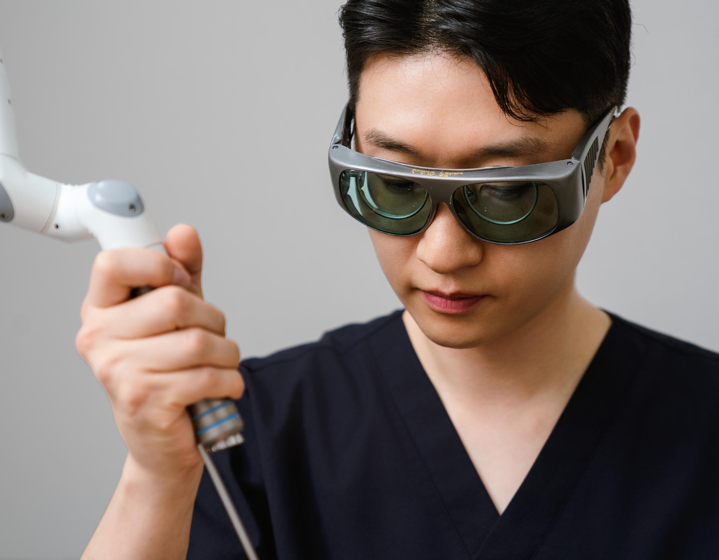 clinician in dark scrubs holding a handheld dermatology laser device, wearing protective eyewear