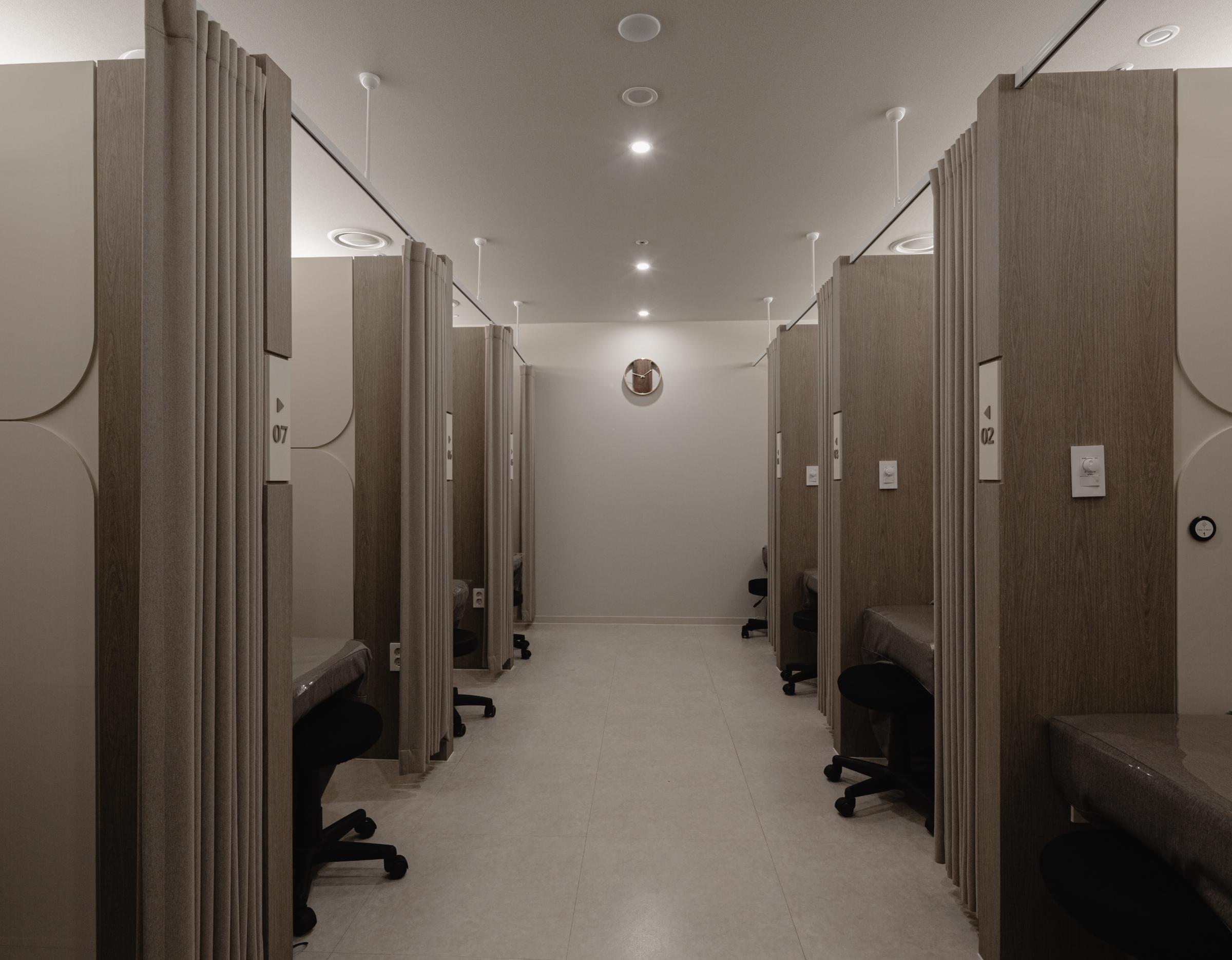 Narrow clinic treatment room corridor with numbered wooden partitioned cubicles, reclining chairs and privacy curtains on both sides