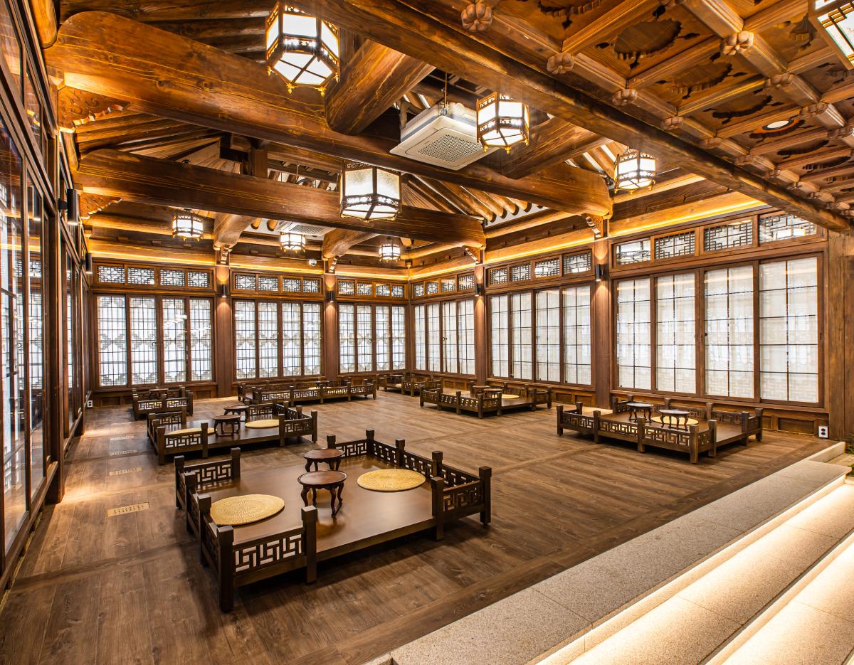 Spacious traditional Korean hanok interior with low wooden tables, floor cushions, intricate beams and lattice windows lit by hanging lanterns