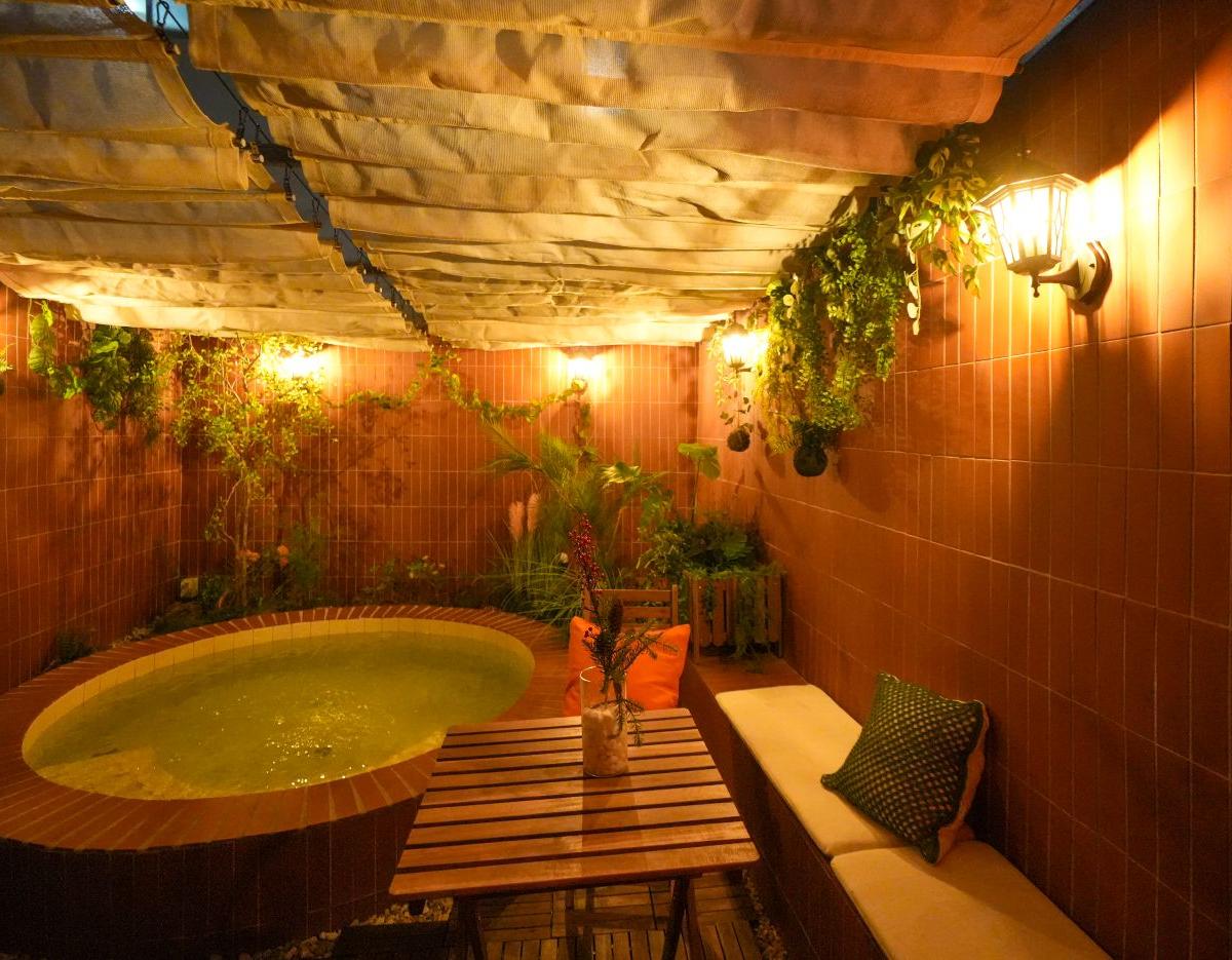 Private indoor jacuzzi niche with round tiled tub, wooden table, cushioned bench and hanging plants under warm wall lamps