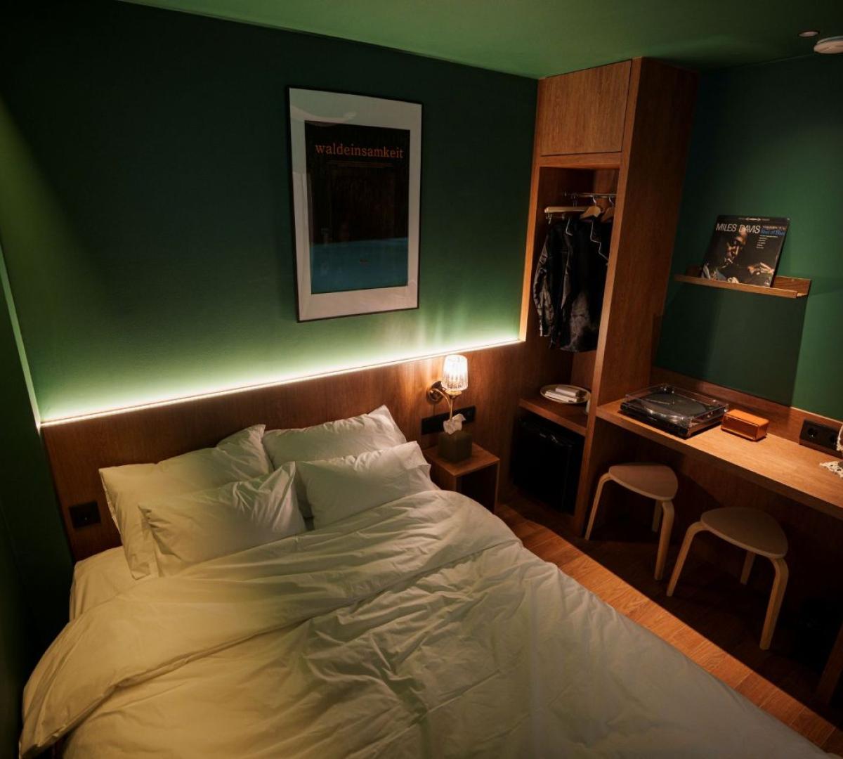 dimly lit green-walled room with a double bed, wooden headboard, open wardrobe with jackets, desk and vinyl player
