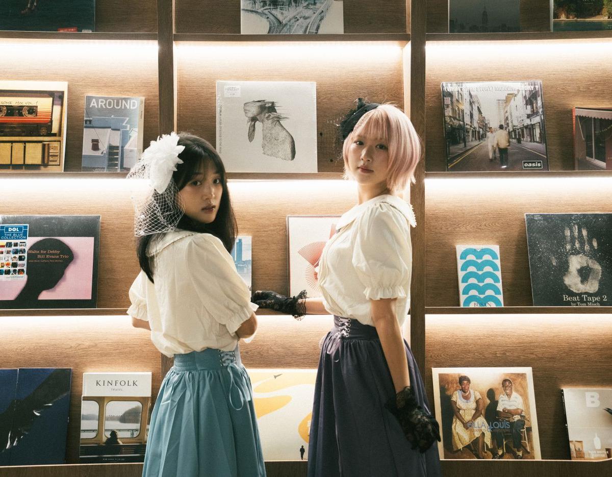 two women in vintage-inspired blouses and waist-cinching skirts pose facing illuminated record-filled wooden shelves, one wearing a short pink wig and lace gloves
