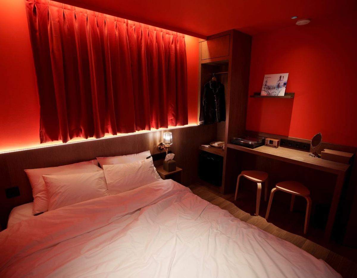 Retro-inspired hotel room with red walls and curtains, low bed with white linens, bedside lamp, open wardrobe and wooden desk with stools.