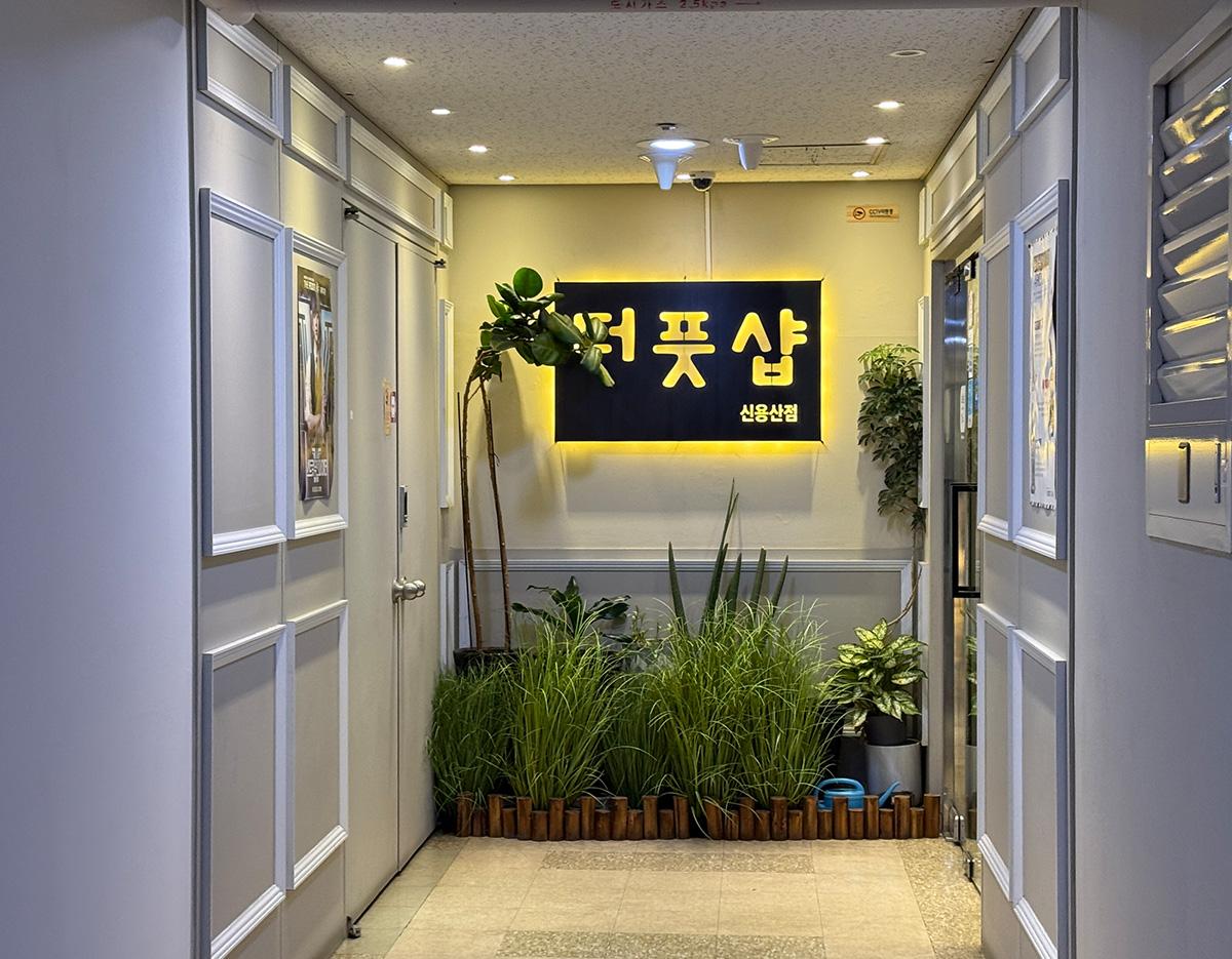 narrow hallway entrance with potted plants and illuminated black-and-yellow sign reading "더 풋샵 신용산점