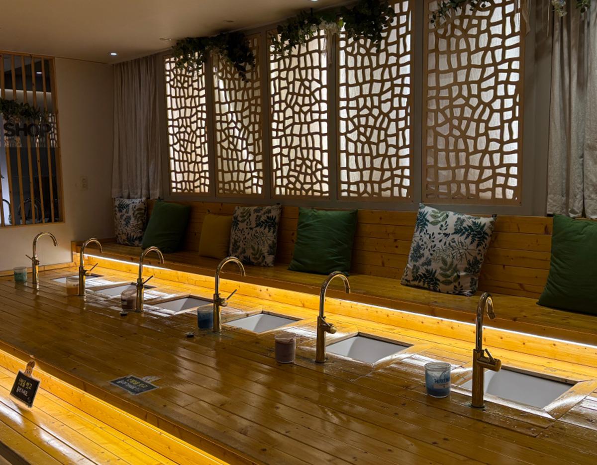 Wooden foot bath bench with multiple faucets, recessed basins and decorative lattice screens with green cushions behind.