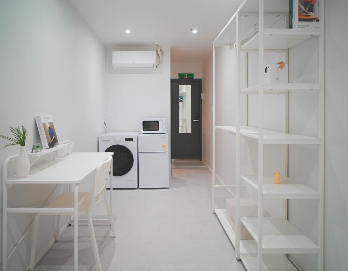 Minimal white single-occupancy room with desk and chair, washer, mini-fridge and microwave, open shelving and air conditioner.