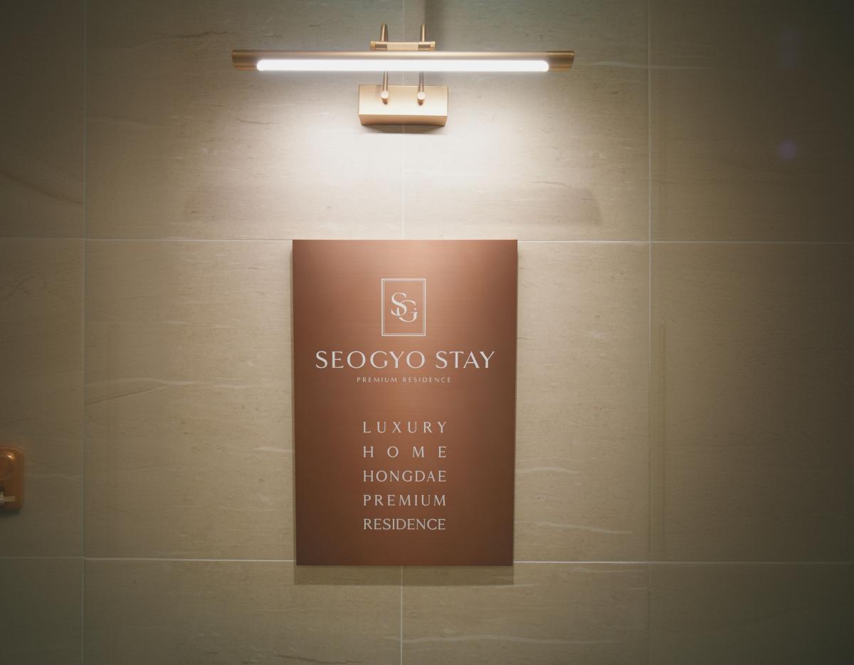 bronze wall plaque reading "SEOGYO STAY" and details for a luxury Hongdae premium residence lit by a brass wall light