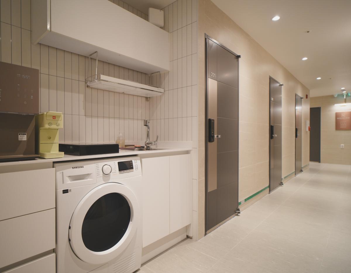 Hallway with multiple locked gray room doors and a white kitchenette area featuring a front-load Samsung washer and sink.
