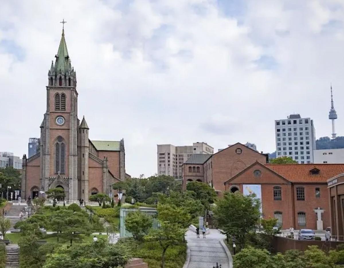 Seoul cityscape with red-brick Myeongdong Cathedral and surrounding greenery, pathways and distant Namsan Tower.