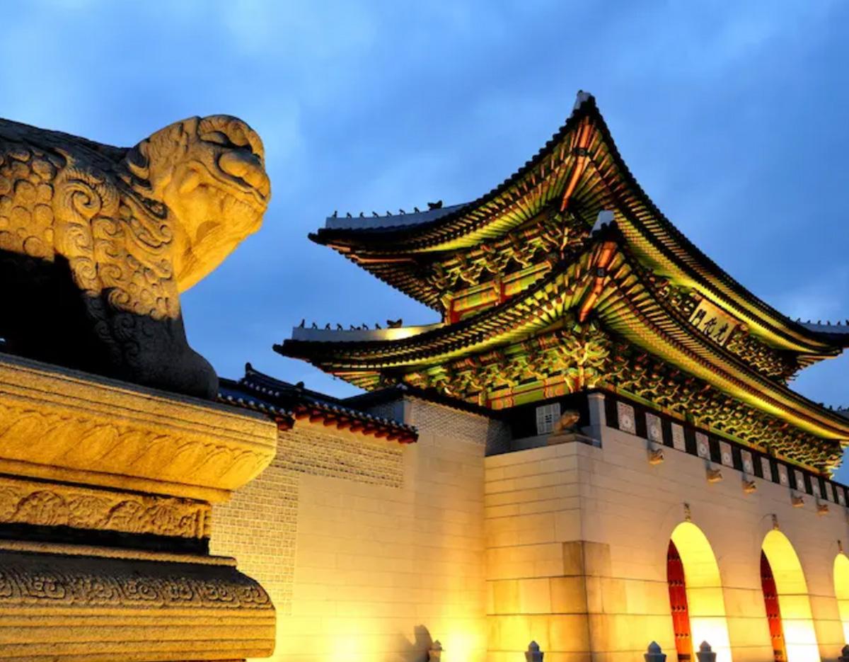 Gyeongbokgung palace gate lit at dusk with ornate multicolored eaves and a carved stone guardian statue in foreground