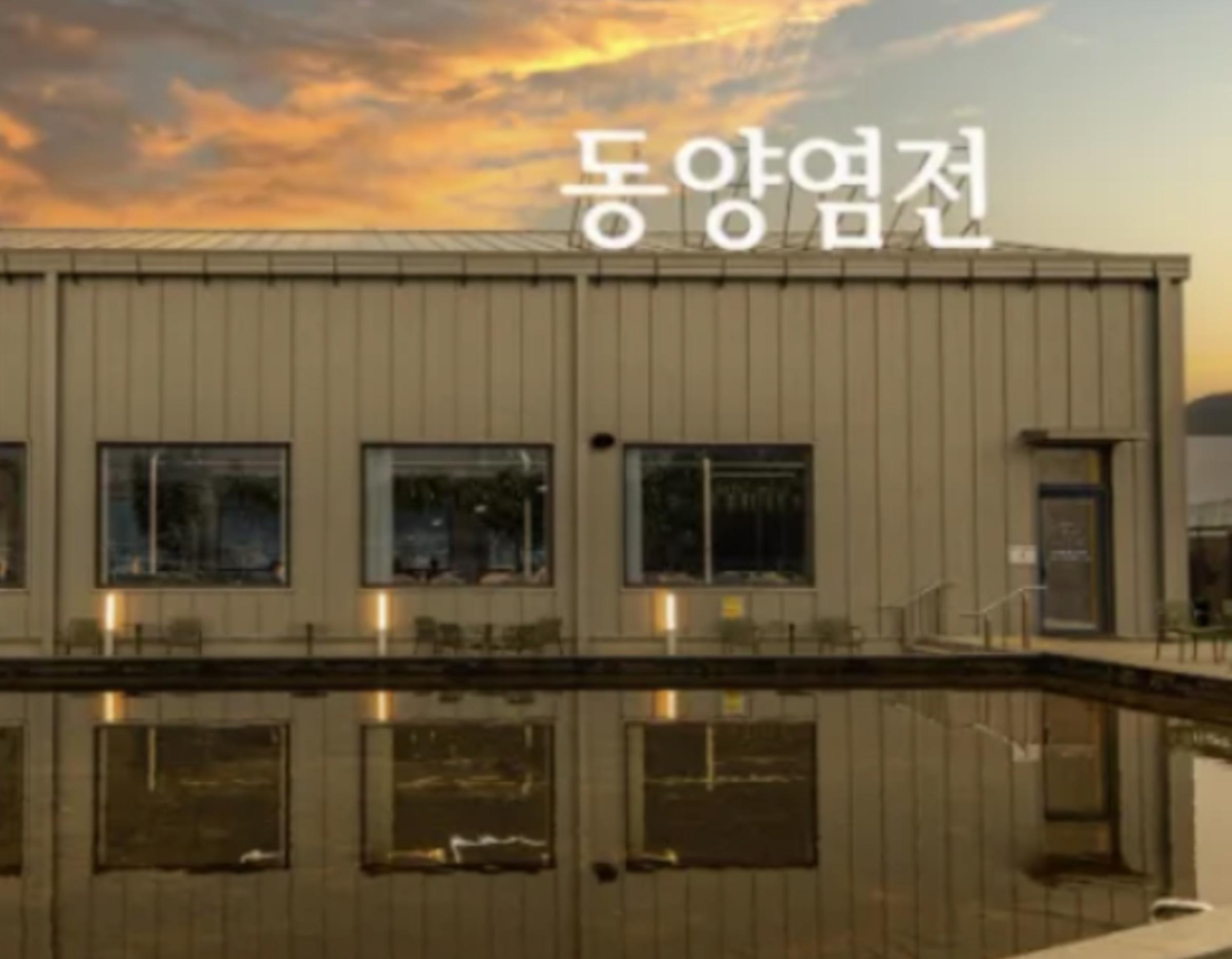 metallic seaside bakery building "동양염전" reflected in still water at sunset with orange clouds above