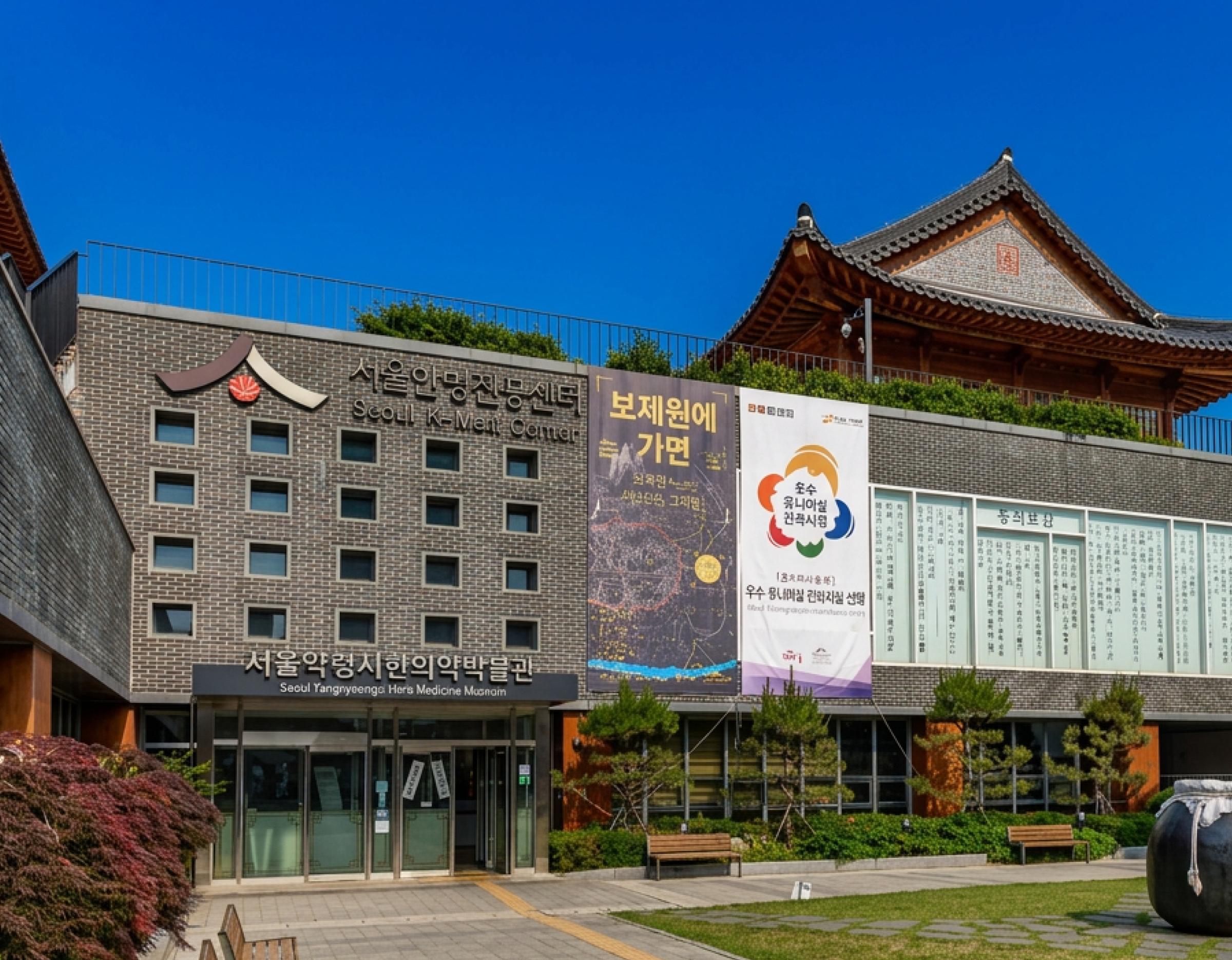 Seoul Hanbang Promotion Center building with traditional tiled pavilion on roof, banners and landscaped courtyard benches