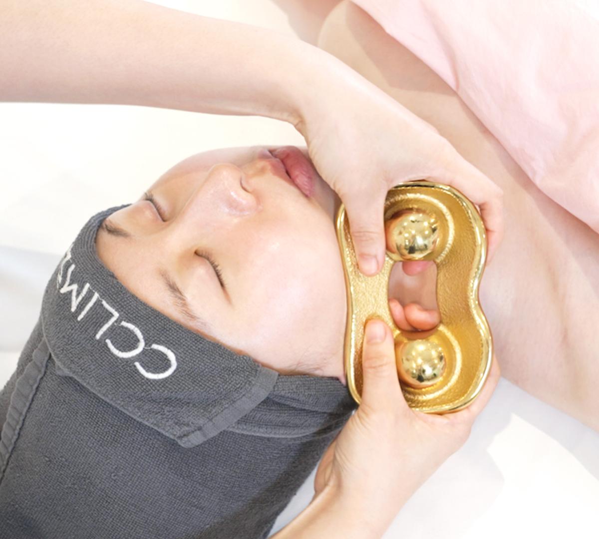 therapist's hands holding a gold dual-ball facial massage tool against a client's jawline on a treatment bed