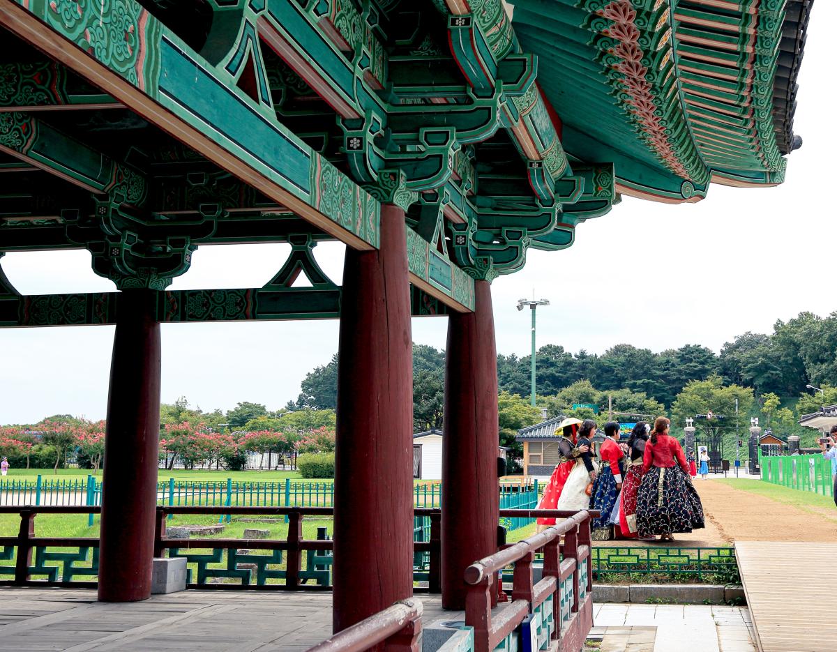 Ornately painted wooden pavilion roof and columns at Gyeongju with visitors in colorful hanbok near the pond