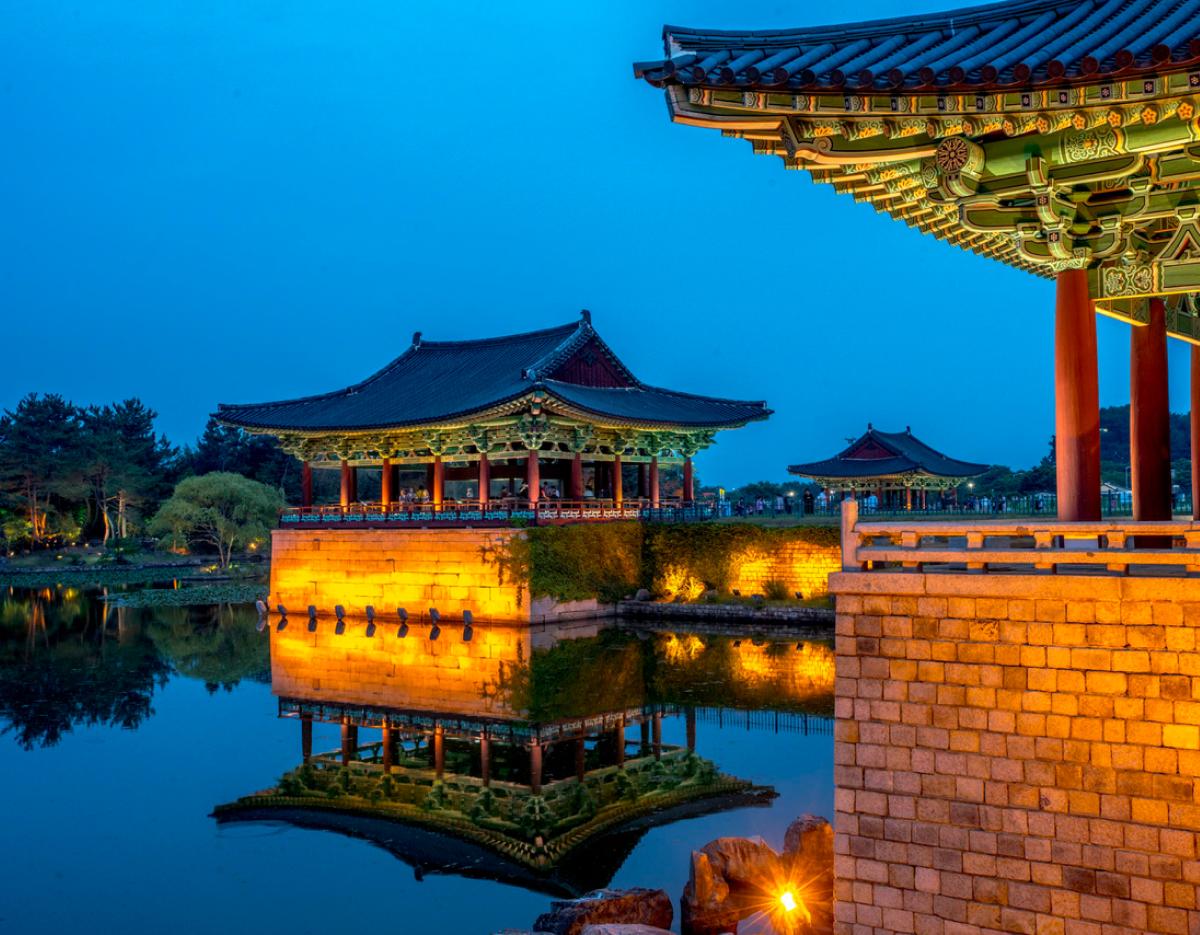 Gyeongju Woljeonggyo/Anapji-style pavilions lit at twilight reflected in calm pond, ornate painted eaves and stone terraces visible
