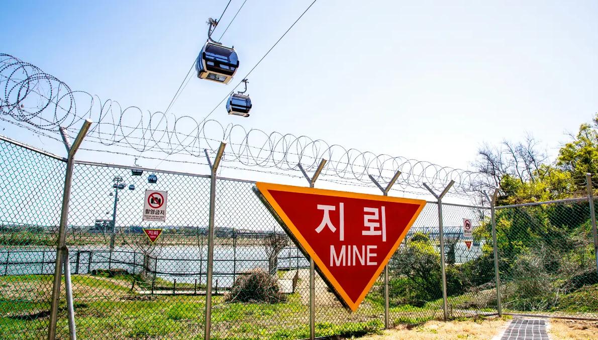 red triangular "MINE" warning sign in front of barbed-wire fence and cable car cabins over the Imjin River
