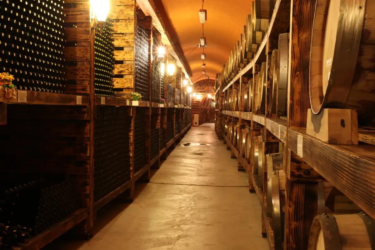 dimly lit winery cellar corridor lined with stacked wine bottles on left and oak barrels on wooden racks right