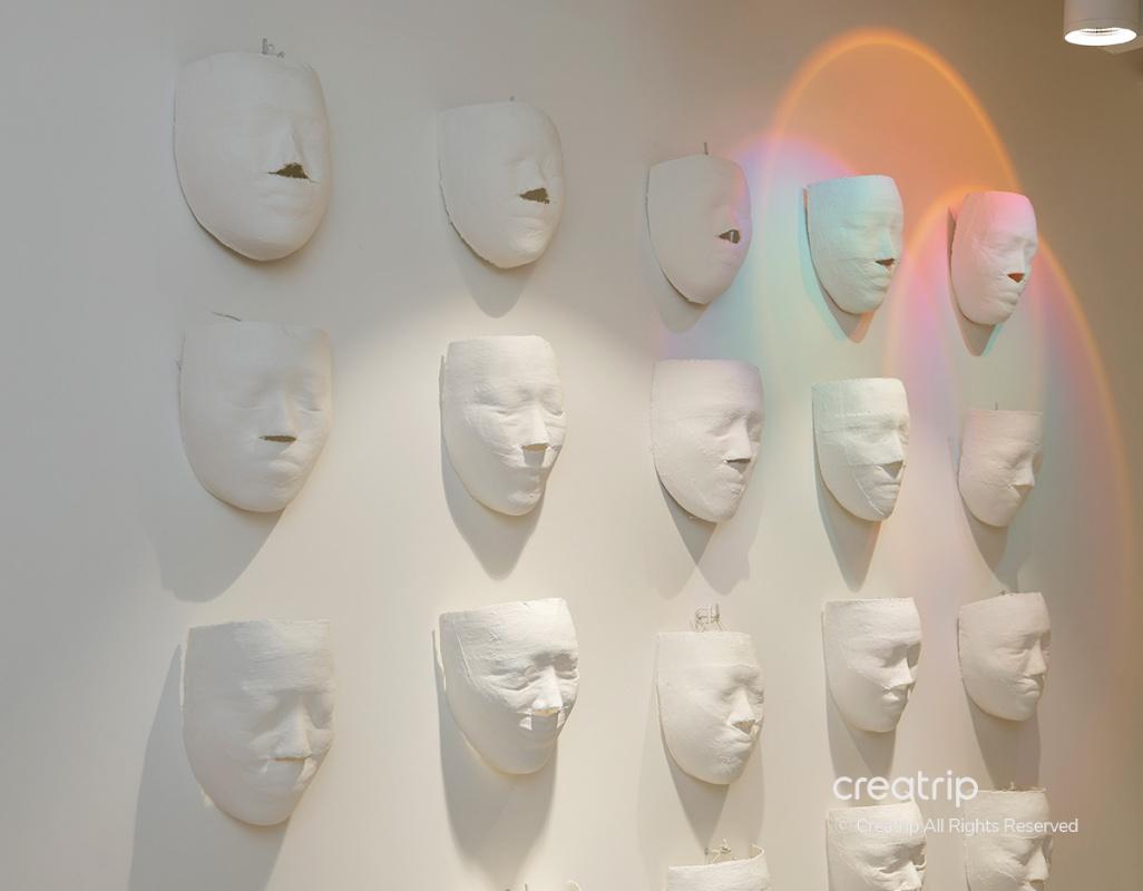 white sculpted face masks arranged in a grid on a cream gallery wall with soft spotlight and rainbow light flare