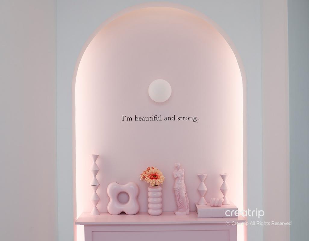 Pastel pink arched niche with decorative vases, sculpture, flowers and wall text reading "I'm beautiful and strong.