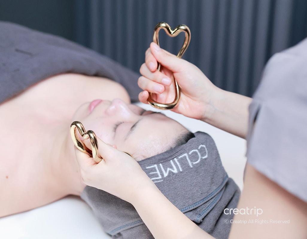 therapist's hands holding a gold dual-ball facial massage tool against a client's jawline on a treatment bed