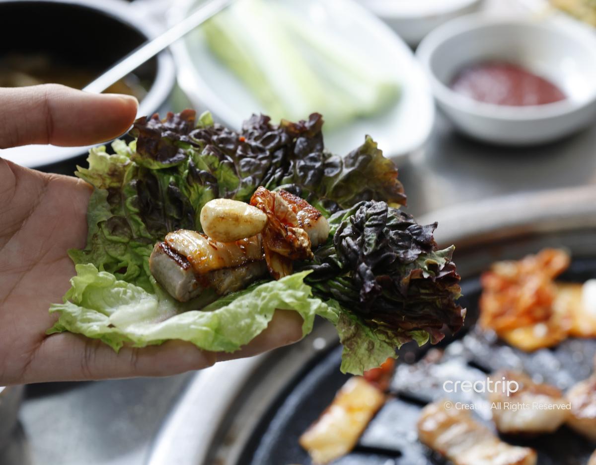 hand holding lettuce wrap with grilled pork belly, garlic and kimchi over a charcoal grill at 청기와갈비 in Hongdae