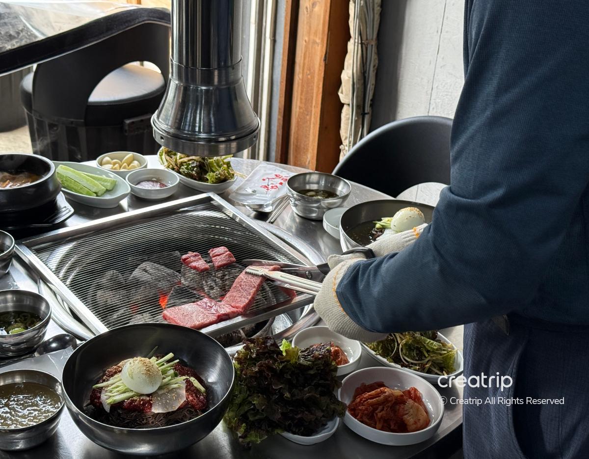 server grilling marbled beef over charcoal grill with banchan bowls, lettuce, and cold noodle bowls at 청기와갈비 table