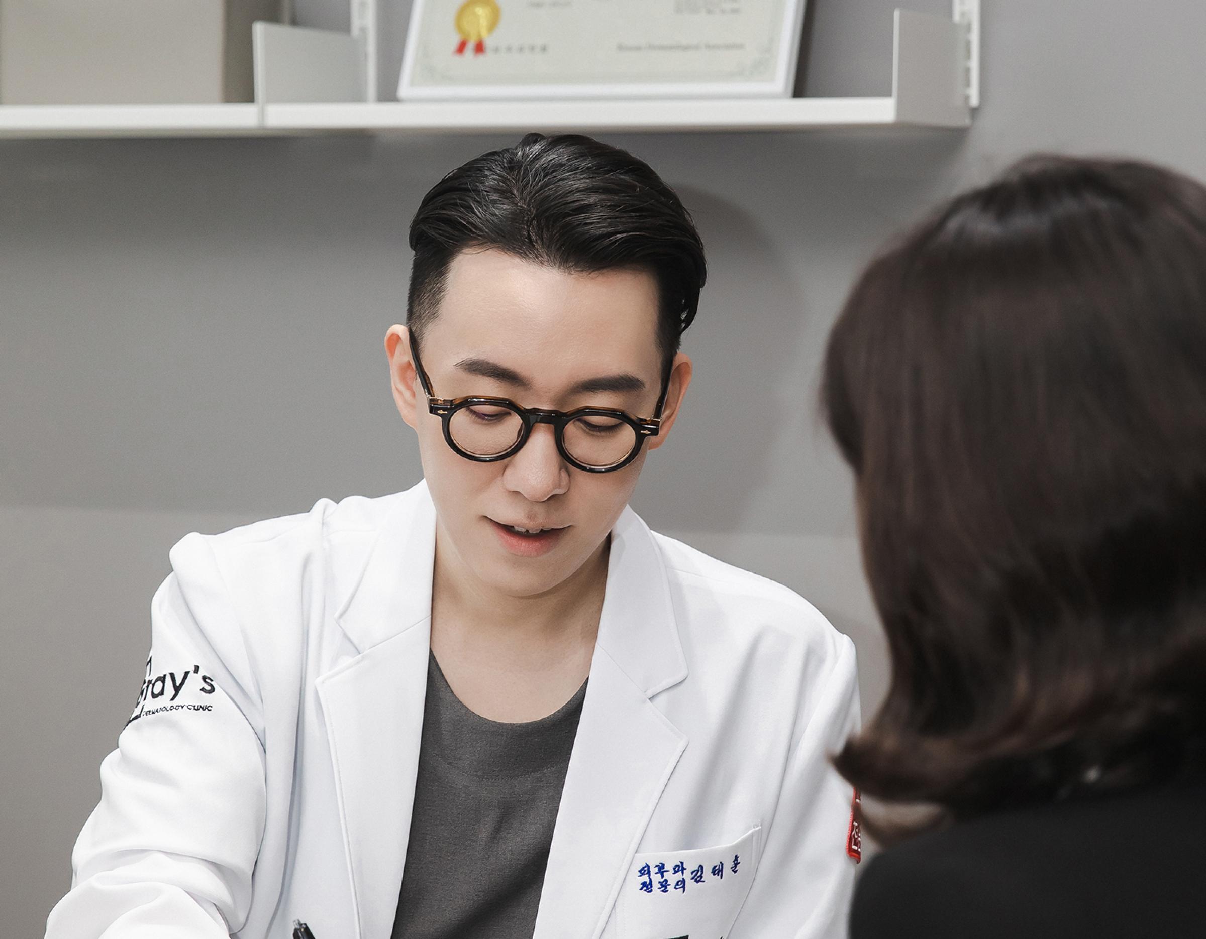 male doctor in white coat consulting seated with patient, framed certificates and shelves in background