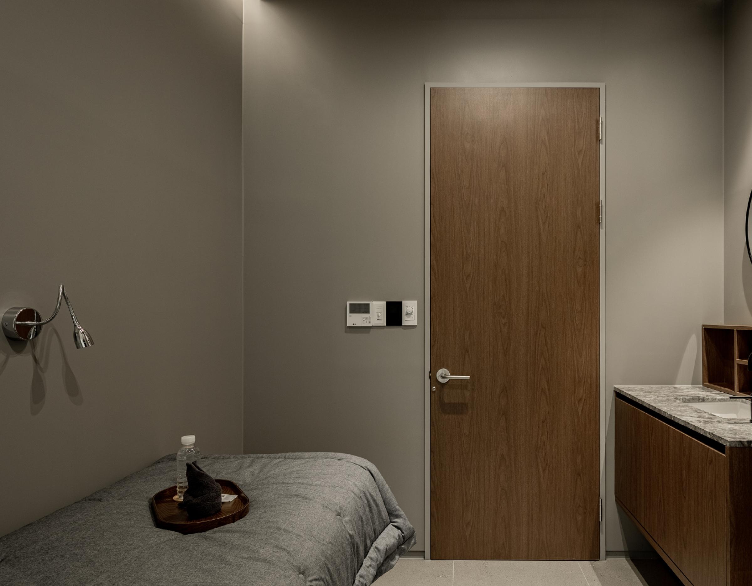 Minimal private clinic treatment room with wooden door, gray treatment bed, tray with water bottle — 그레이스피부과.