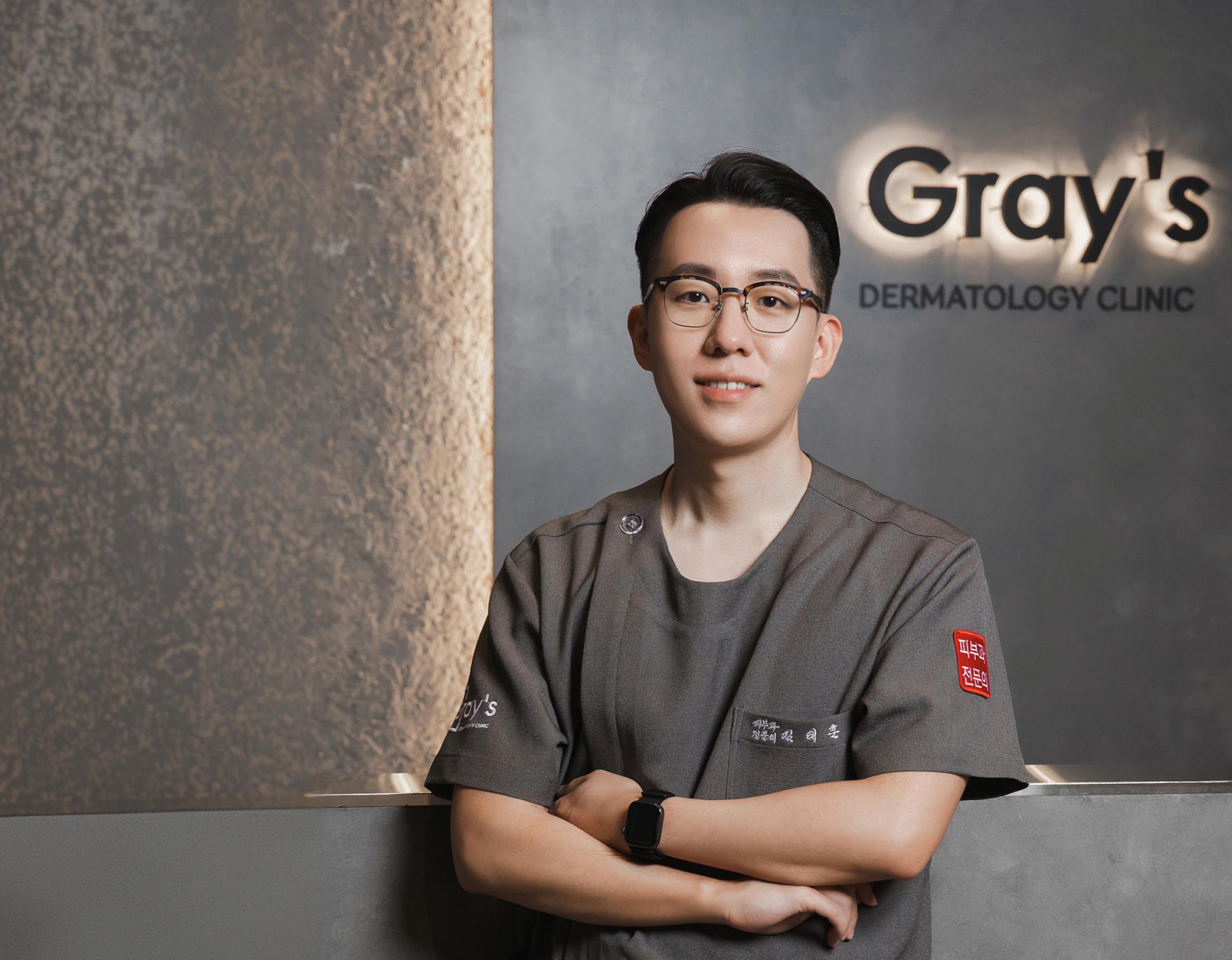 Male clinician with blurred face standing arms crossed in gray scrub top in front of backlit "Gray's Dermatology Clinic" sign.