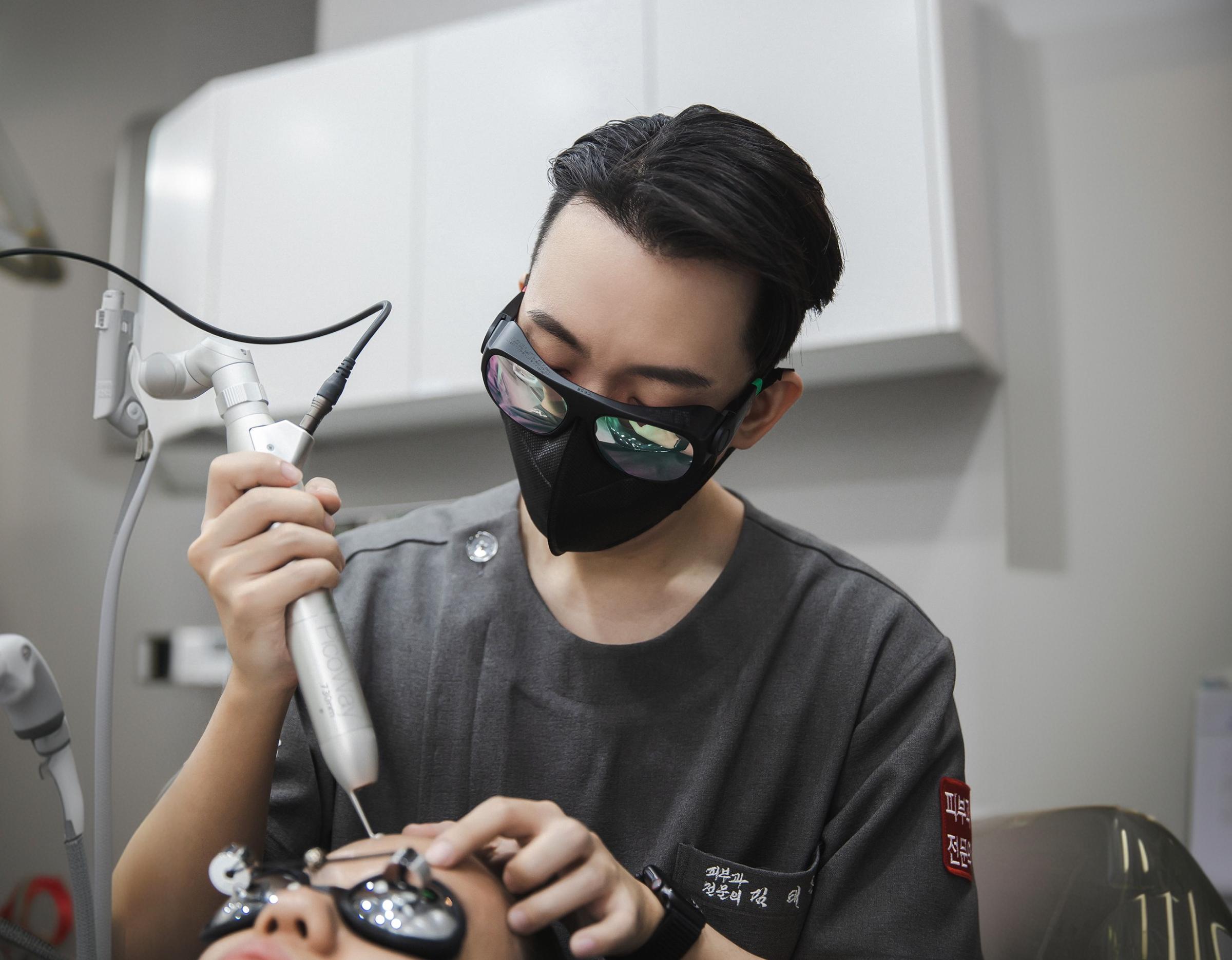 Dermatologist holding a handheld laser device over a reclining patient's face wearing protective eye shields in a clinic room