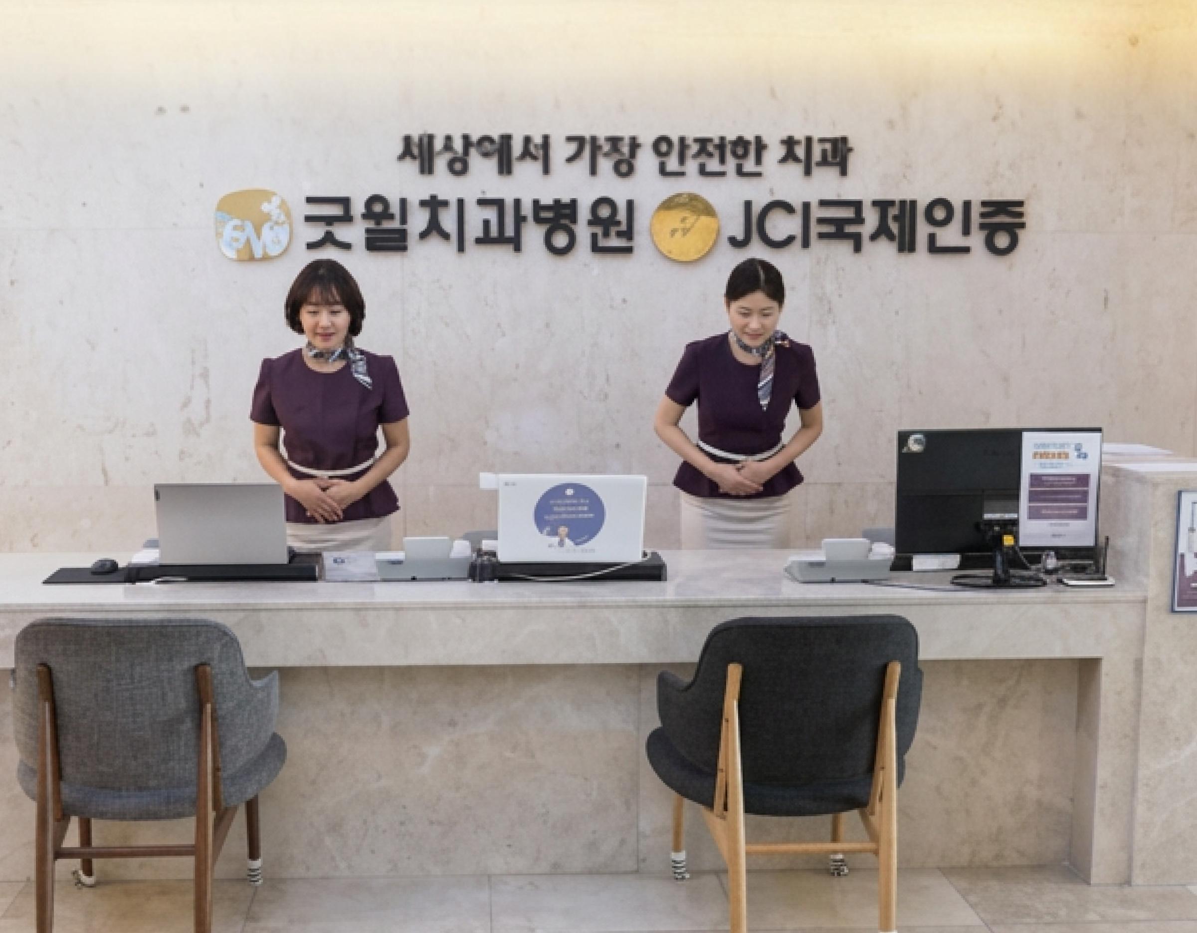 Reception desk with two uniformed staff bowing in front of a marble wall displaying 굿윌치과병원 and JCI 국제인증 signage.